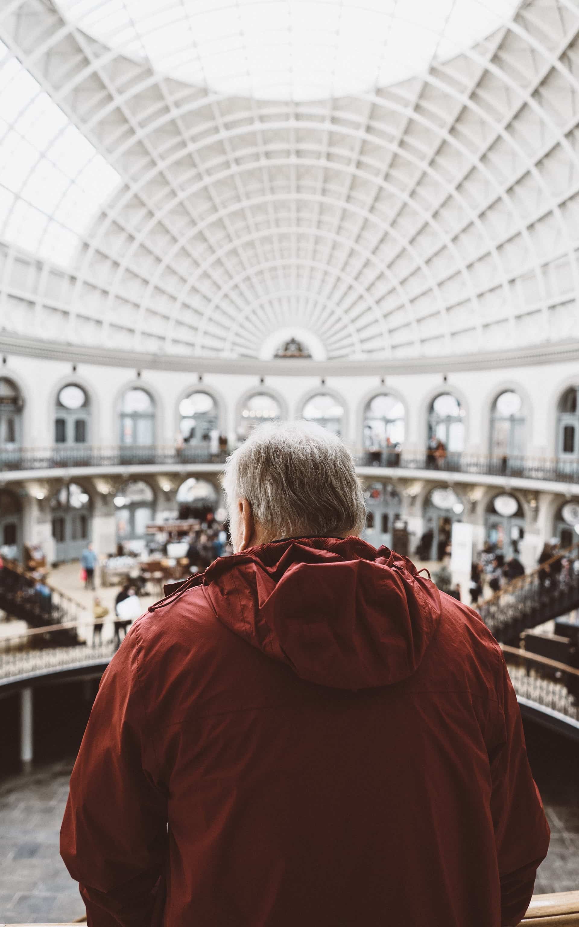 An old man in a red jacket. He is looking out on a hall full of people.